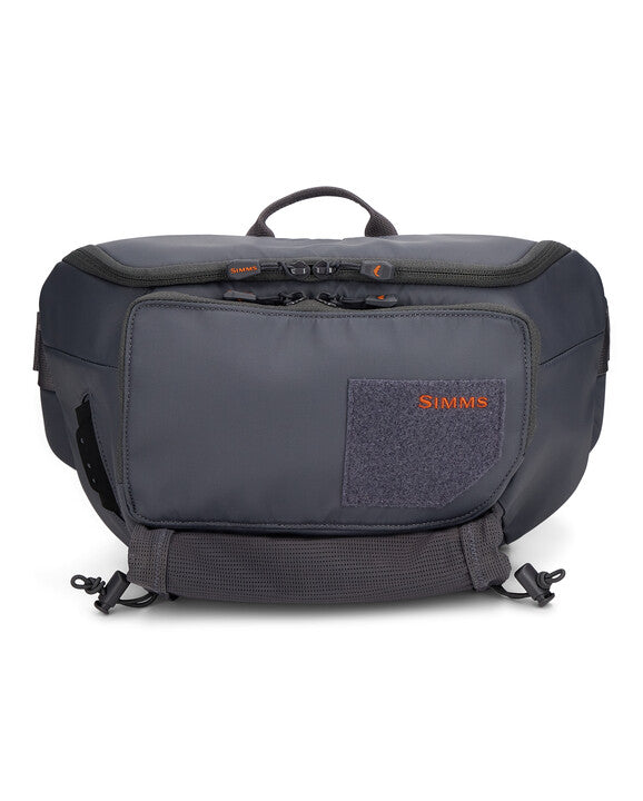 Simms Tributary Hip Pack in dark gray with multiple zippered compartments, adjustable waist strap, and a small orange logo, designed for organizing fishing tackle and gear.