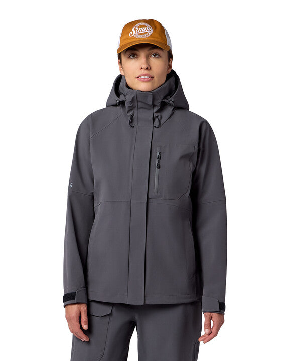 Woman wearing a dark gray Simms W's Challenger Rain Jacket with a hood, zippered chest pocket, and adjustable wrist straps, styled for outdoor fishing activities.