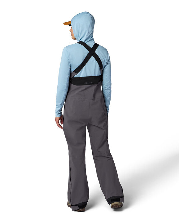 Rear view of a person wearing Simms W's Challenger Rain Bib in gray with black adjustable suspenders and a light blue hooded jacket, designed for water-resistant protection during fishing.