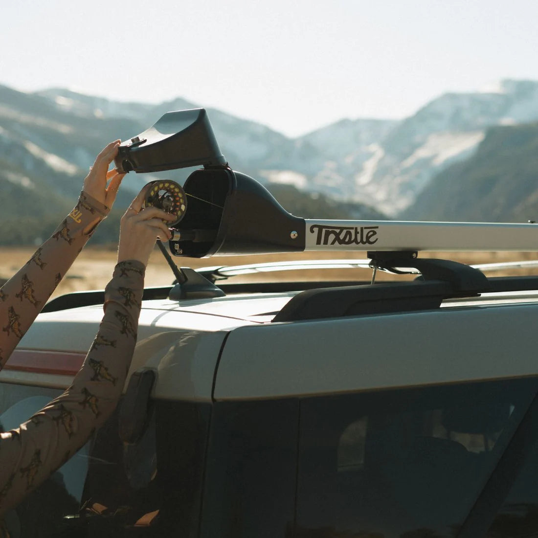 Trxstle CRC Flr Rod Carrier V3.0 mounted on a vehicle roof rack with a fishing rod carrier being secured against a scenic mountain backdrop