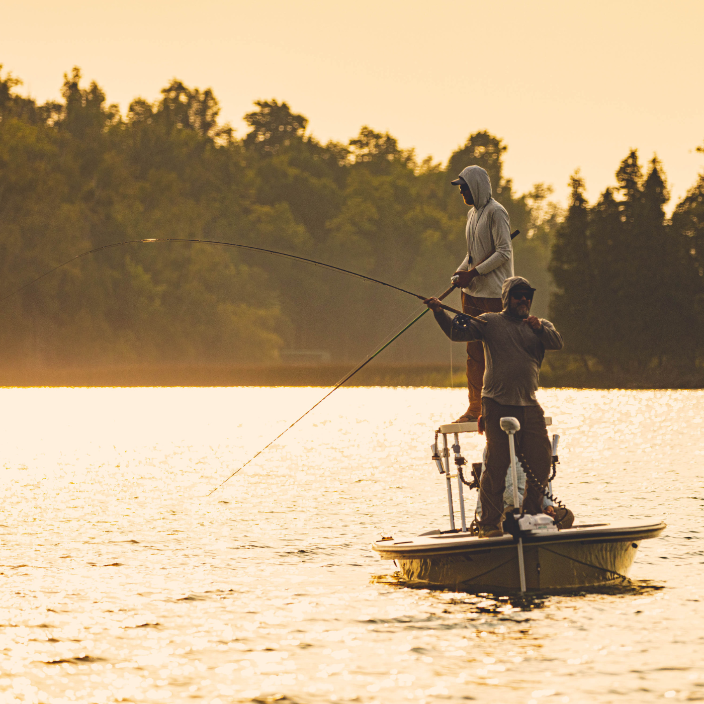 Two fishermen on a small boat casting lines at sunset using the Thorne Bros Musky Fly Rod - Heavy in a serene lake setting surrounded by trees.