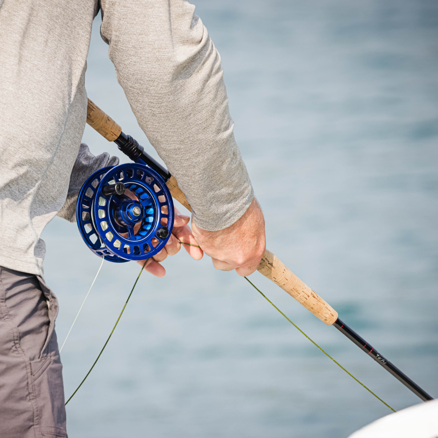 Angled close-up of a person holding a Thorne Bros Musky Fly Rod - Heavy with cork handle, detailed blue reel, and fishing line over water background.