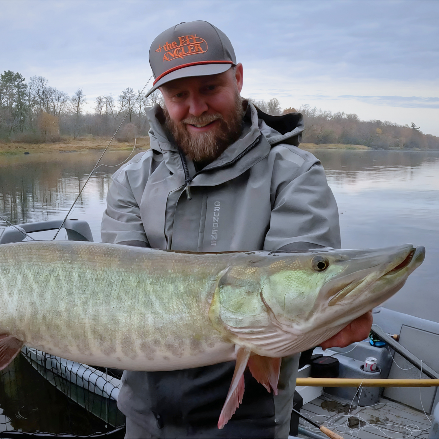 Man wearing a gray jacket and hat smiling while holding a large muskellunge fish on a boat, highlighting the use of the Thorne Bros Musky Fly Rod - Heavy for musky fishing.
