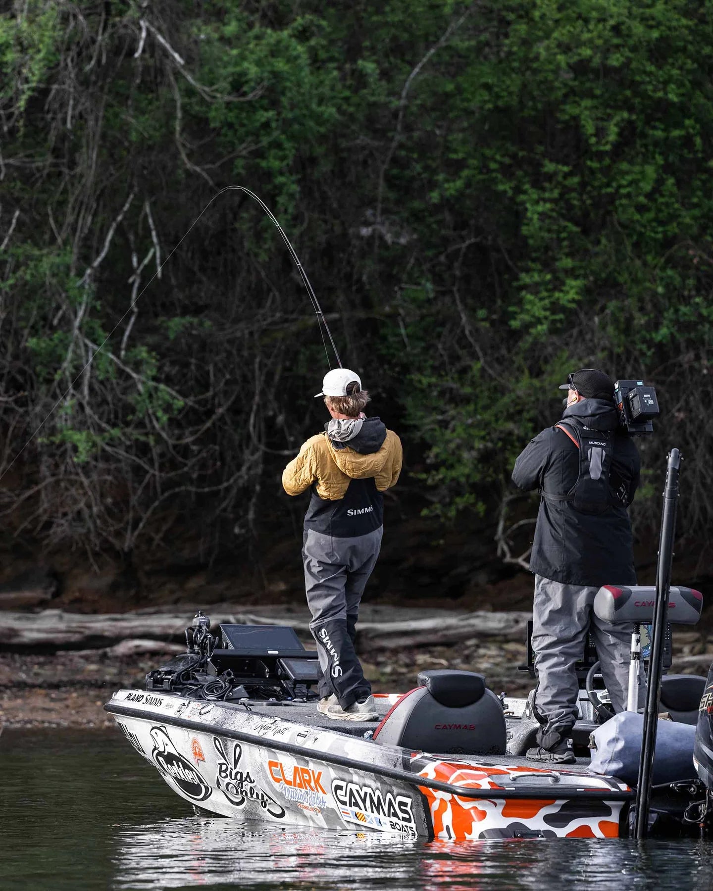 Man fishing on a boat wearing Simms Men's Challenger Rain Bib-Jet Black, casting a fishing rod near a wooded shoreline while being filmed by a cameraman.