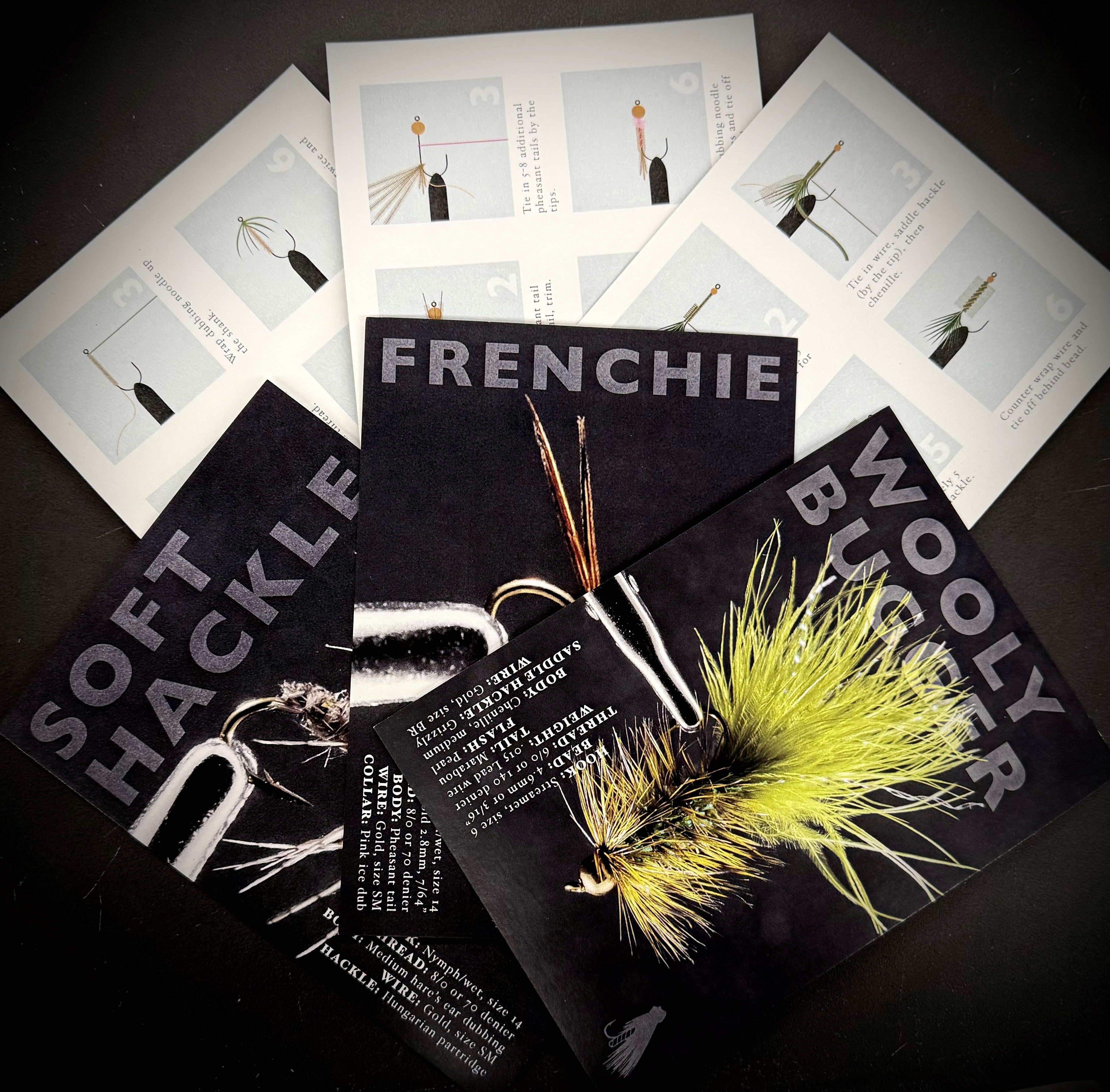 Fly Tying Class - Beginner materials including instructional cards and examples of fly fishing lures such as Frenchie, Soft Hackle, and Woolly Bugger displayed on a dark surface.