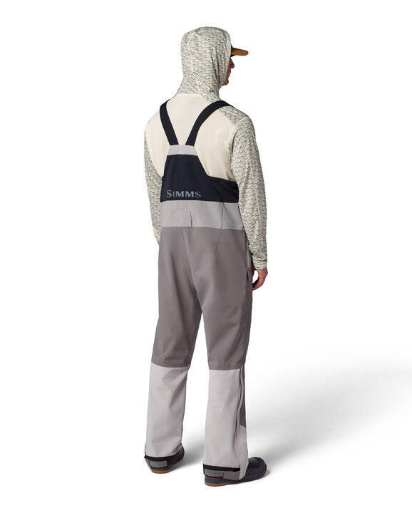 Simms Men's Challenger Rain Bib shown from the back, featuring adjustable shoulder straps, durable two-tone gray waterproof fabric, and waterproof boots for fishing and outdoor protection.