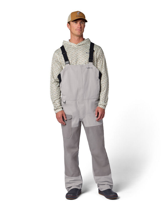 Man wearing light gray Simms Men's Challenger Rain Bib with black adjustable shoulder straps, paired with a patterned hoodie, tan cap, and black shoes.