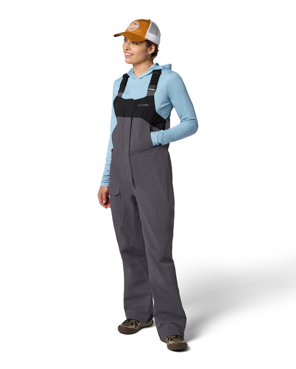 Woman wearing gray and black Simms W's Challenger Rain Bib with adjustable black shoulder straps, paired with light blue hoodie and brown cap, standing in relaxed pose.