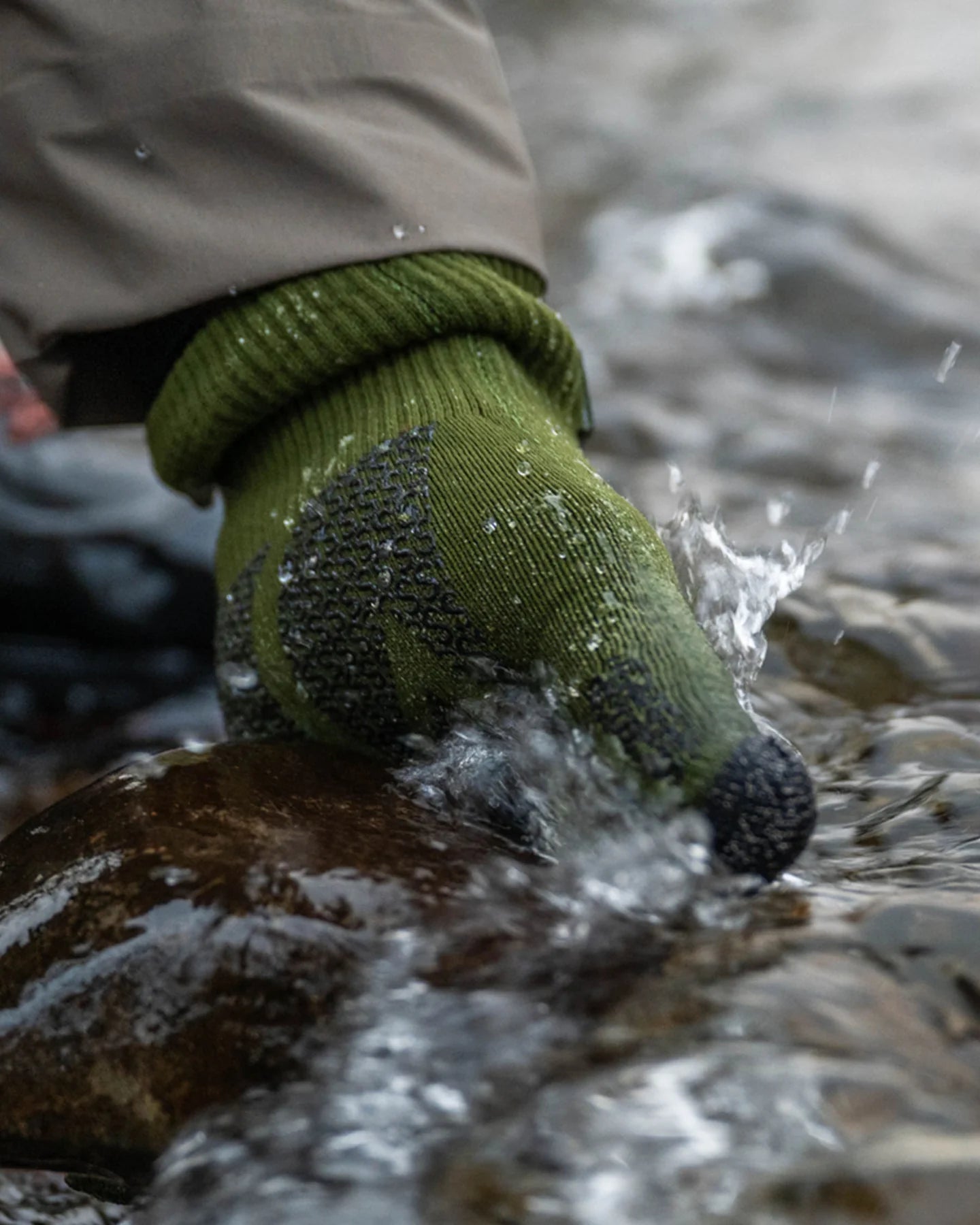 Close-up of a hand wearing a green Simms X Sealskinz Rangeley Waterproof Glove, partially submerged in flowing water, demonstrating its water-resistant and textured grip features.