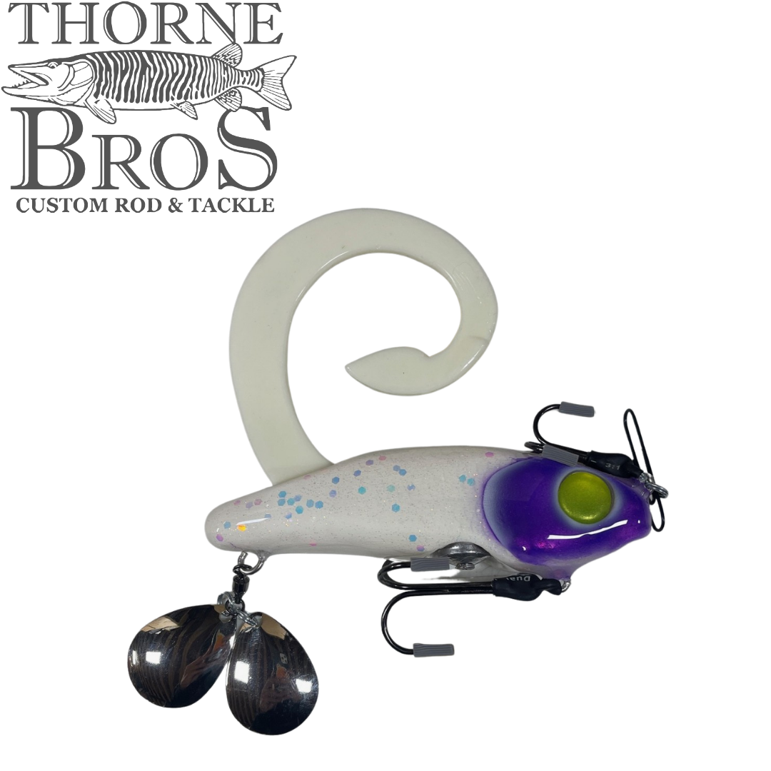 Zilla Baits Gladiator (Mini & Standard) fishing lure featuring a white body with blue speckles, a purple head with yellow eyes, a curled tail, and dual metal spinner blades.