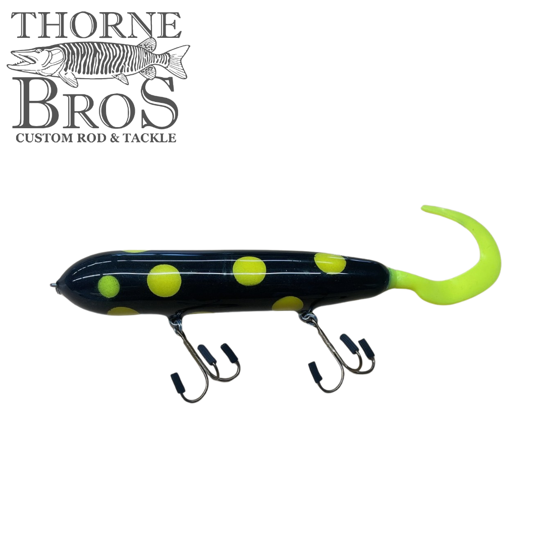 Holmgren Serpent Musky Top Water fishing lure in black with large yellow spots and a bright yellow curved tail, equipped with two treble hooks for musky fishing.