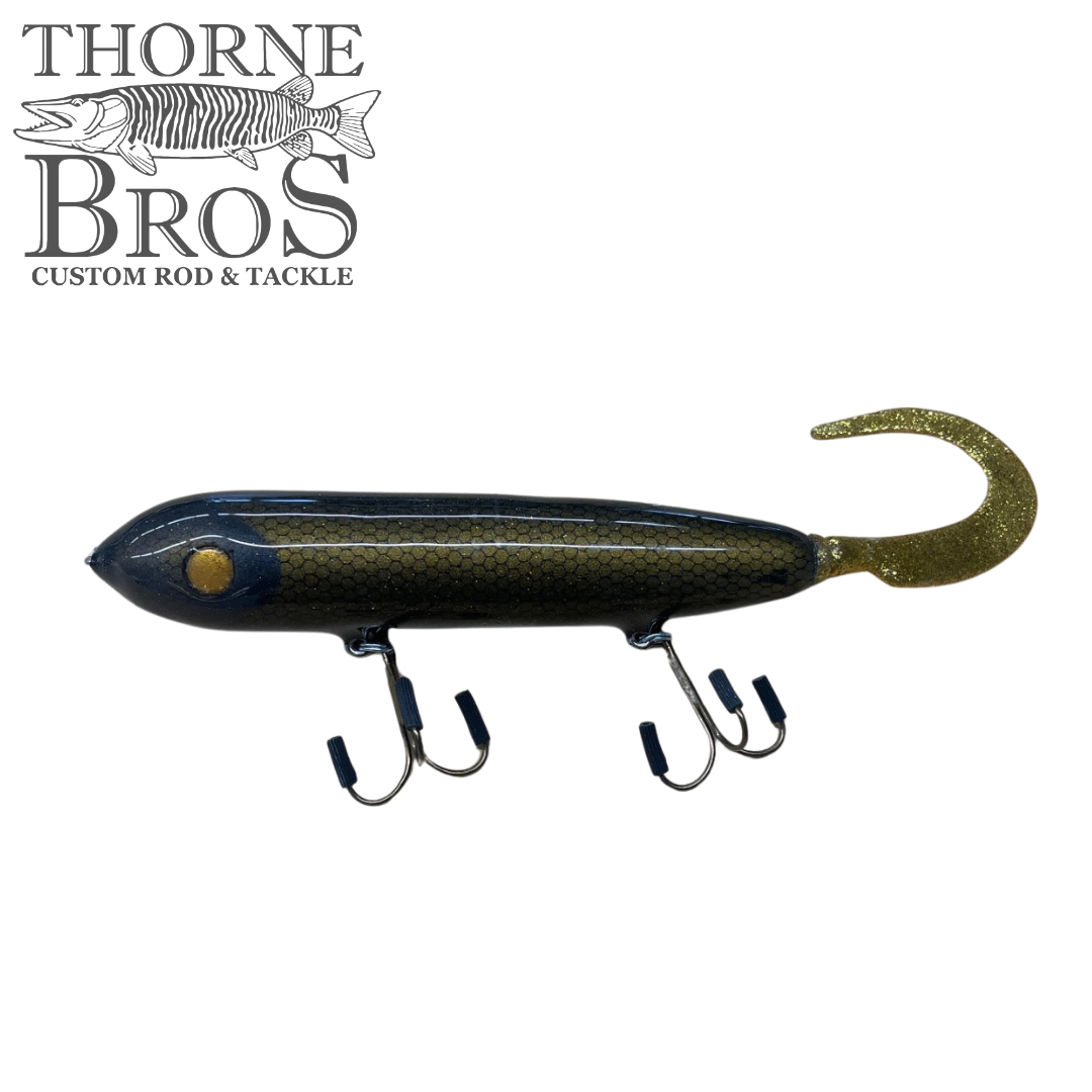 Holmgren Serpent Musky Top Water fishing lure with a dark blue and gold body, featuring two treble hooks and a curly tail designed for musky fishing.