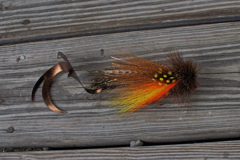 Single Buford Dragon Tail Urban Fly Co. musky fishing lure with orange yellow black and brown feathers and twin curling tails on weathered wood surface